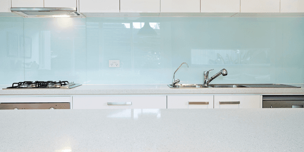 Light blue tinted glass splashback spanning the full width of a modern white kitchen behind a gas cooktop and sink with chrome tapware.