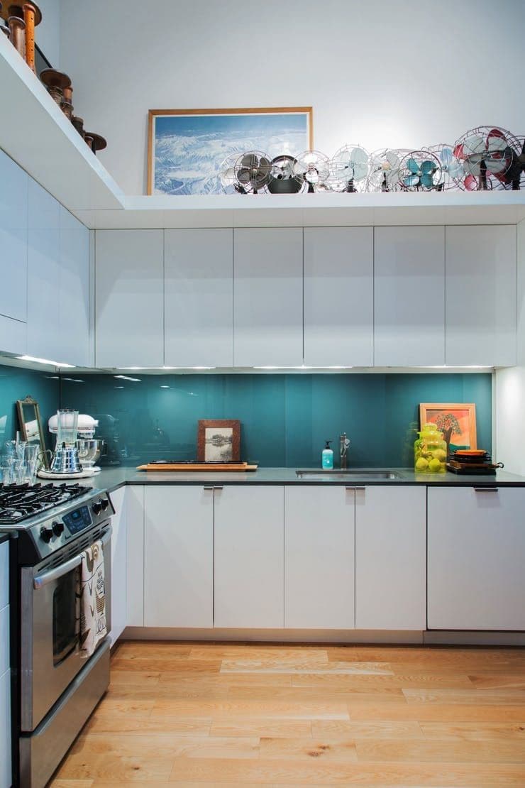 Teal/turquoise coloured glass splashback mounted flush on the wall behind a gas cooktop and stove in a modern white kitchen with dark benchtops.