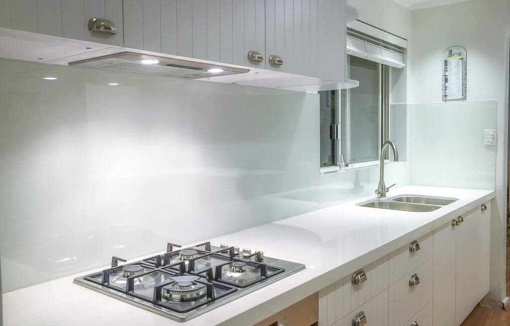 White glass splashback mounted flush on the wall behind a stainless steel gas cooktop and double sink in a bright white Hampton-style kitchen.
