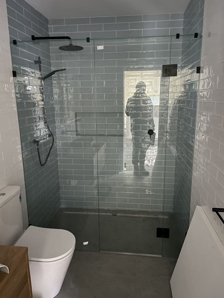 Semi-frameless shower enclosure with a sliding door on a top rail and matte black hardware, featuring clear glass in an L-configuration against grey subway tiled walls with concrete-look floor tiles.