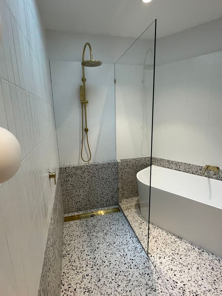 Frameless fixed shower screen panel with brushed gold fittings, separating a wet shower area from a freestanding bathtub in a luxurious terrazzo-floored bathroom.