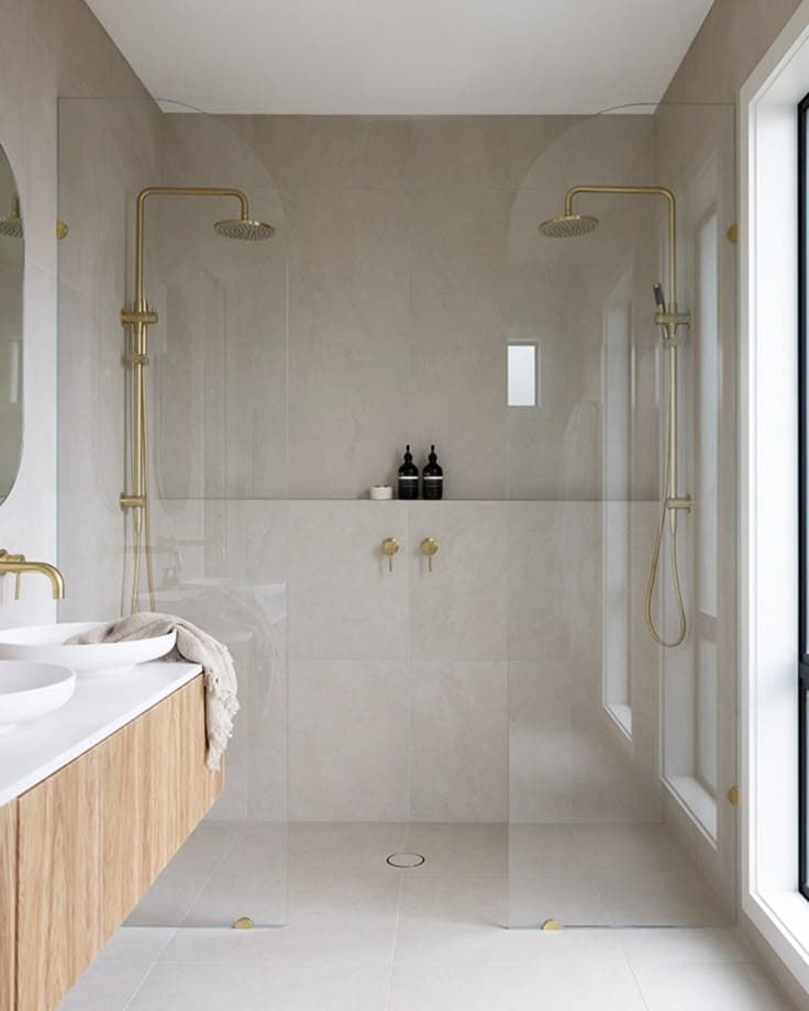 Frameless shower enclosure with brushed gold square hinges and clips mounted directly onto clear glass panels, forming a walk-in configuration in a neutral stone-tiled ensuite with dual brushed gold rainfall shower heads.