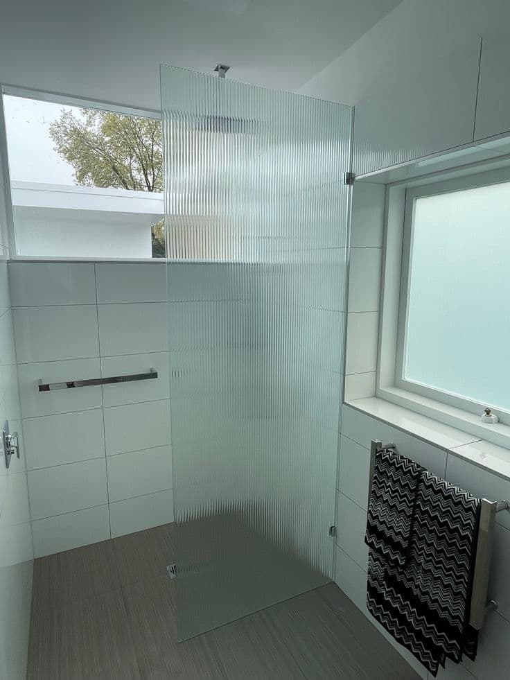 Frameless shower screen featuring reeded glass in a fixed panel configuration with a ceiling-mounted chrome spigot and chrome hardware in a white tiled ensuite.