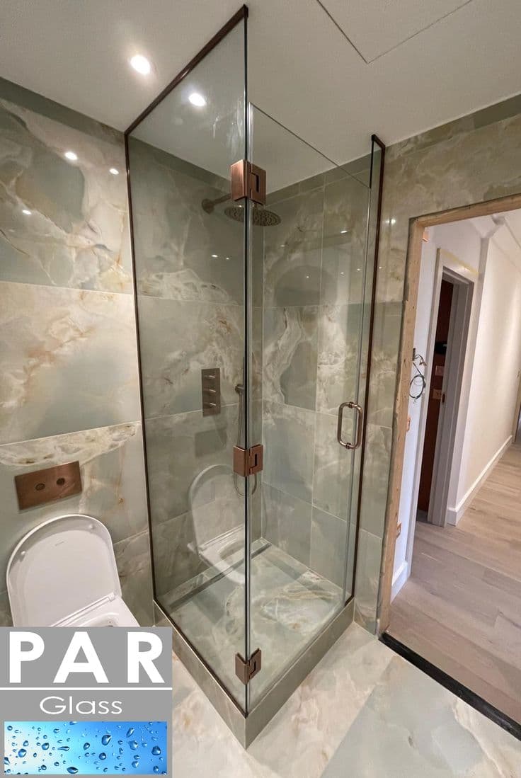 Frameless shower enclosure in a corner configuration with hinges and clips mounted directly onto the glass edges in a brushed gold/copper finish, set in a luxurious marble-tiled bathroom.