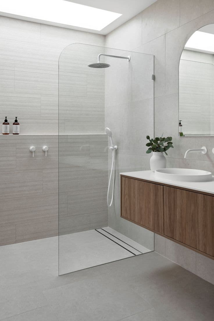 Frameless fixed shower screen with gunmetal hardware clips on a single-panel walk-in configuration in a modern light-tiled bathroom with timber vanity.