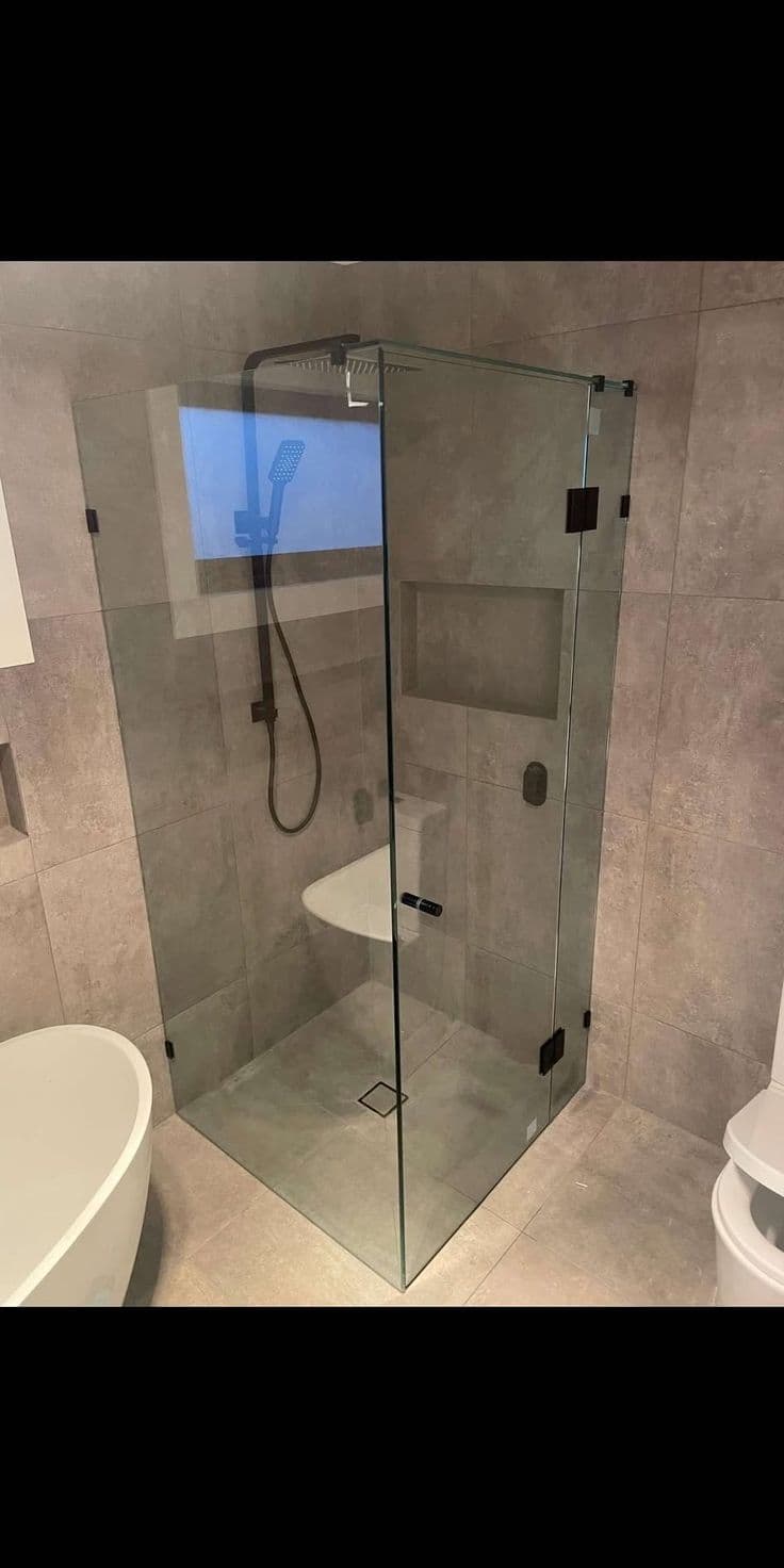 Frameless shower enclosure in a corner configuration with clear glass panels, matte black square hinges and door handle, installed in a modern grey-tiled bathroom with a built-in bench seat.