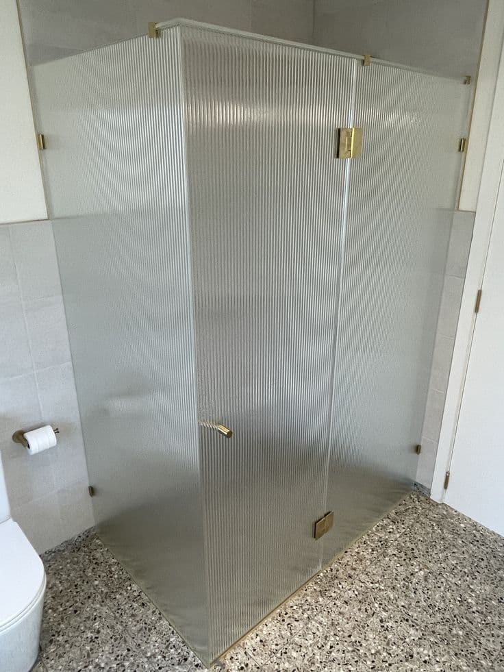 Frameless shower enclosure with reeded glass panels and brushed gold hinges and hardware in a corner configuration with terrazzo flooring.