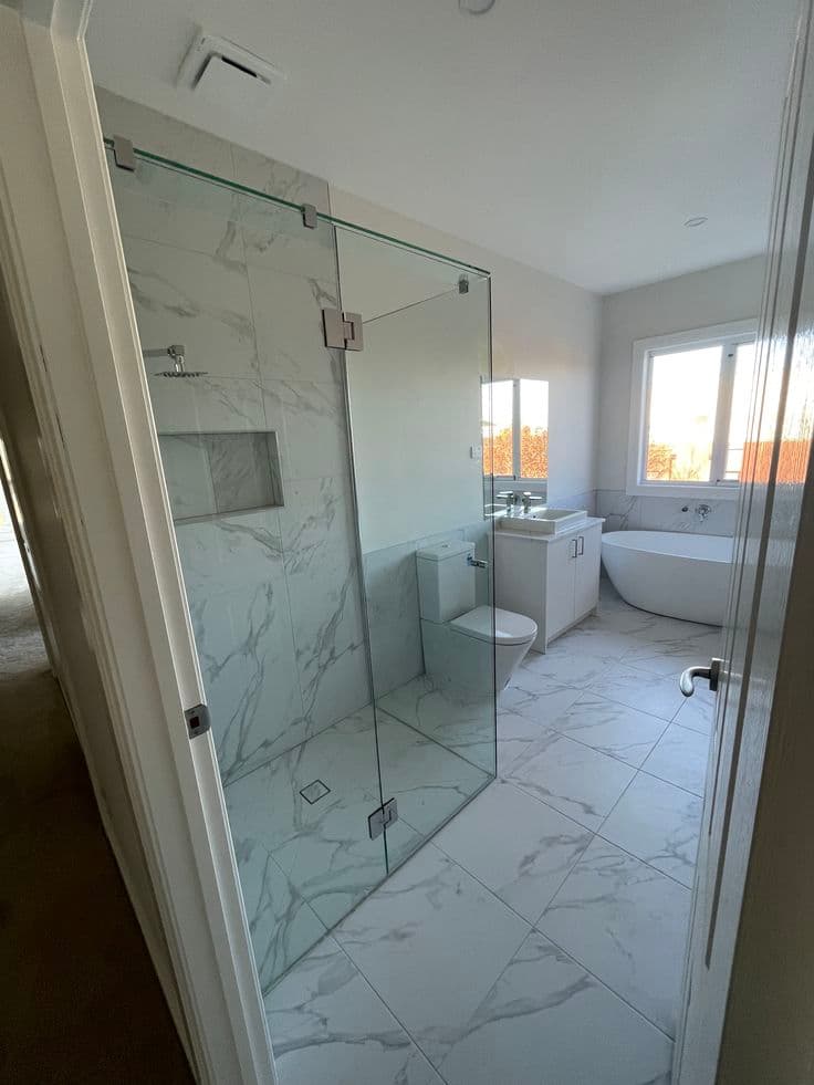 Frameless shower enclosure with chrome hinges and floor-to-ceiling clear glass panels in a corner configuration, set in a luxurious marble-tiled ensuite with freestanding bath.