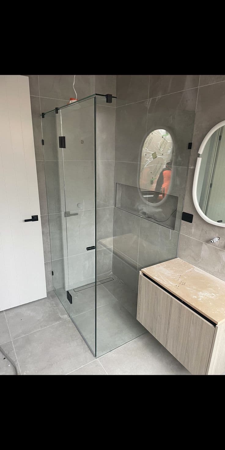 Frameless shower enclosure in a corner configuration with matte black hinges and clips mounted directly onto clear glass panels, set in a modern grey-tiled bathroom with a floating timber vanity.