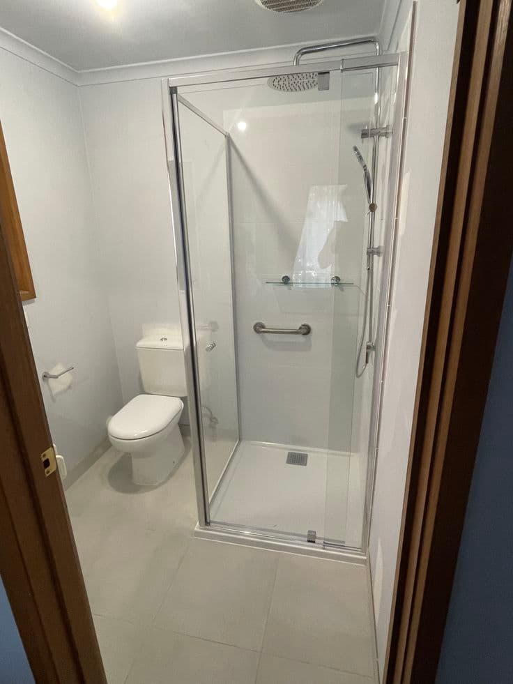 Semi-frameless shower enclosure with chrome aluminium frame rails and pivot door in a compact white tiled bathroom with toilet visible.