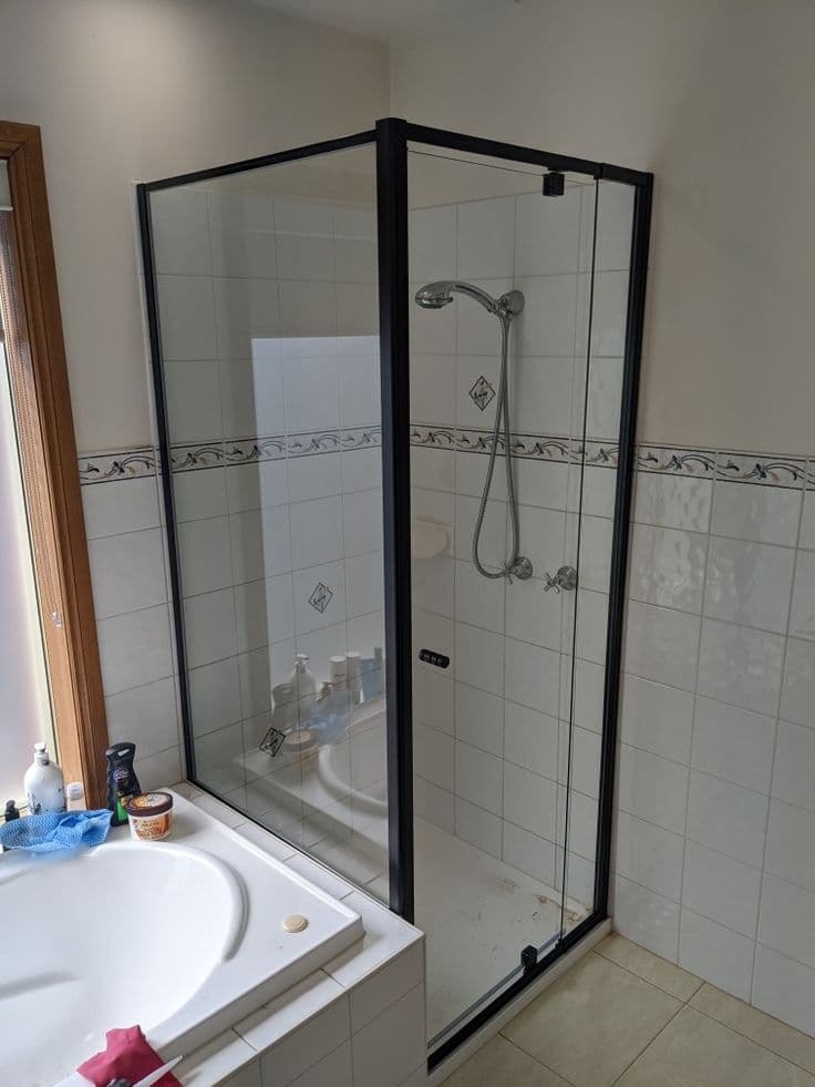 Semi-frameless shower enclosure with matte black aluminium frame rails and corner configuration in a white tiled bathroom with a freestanding bath adjacent.