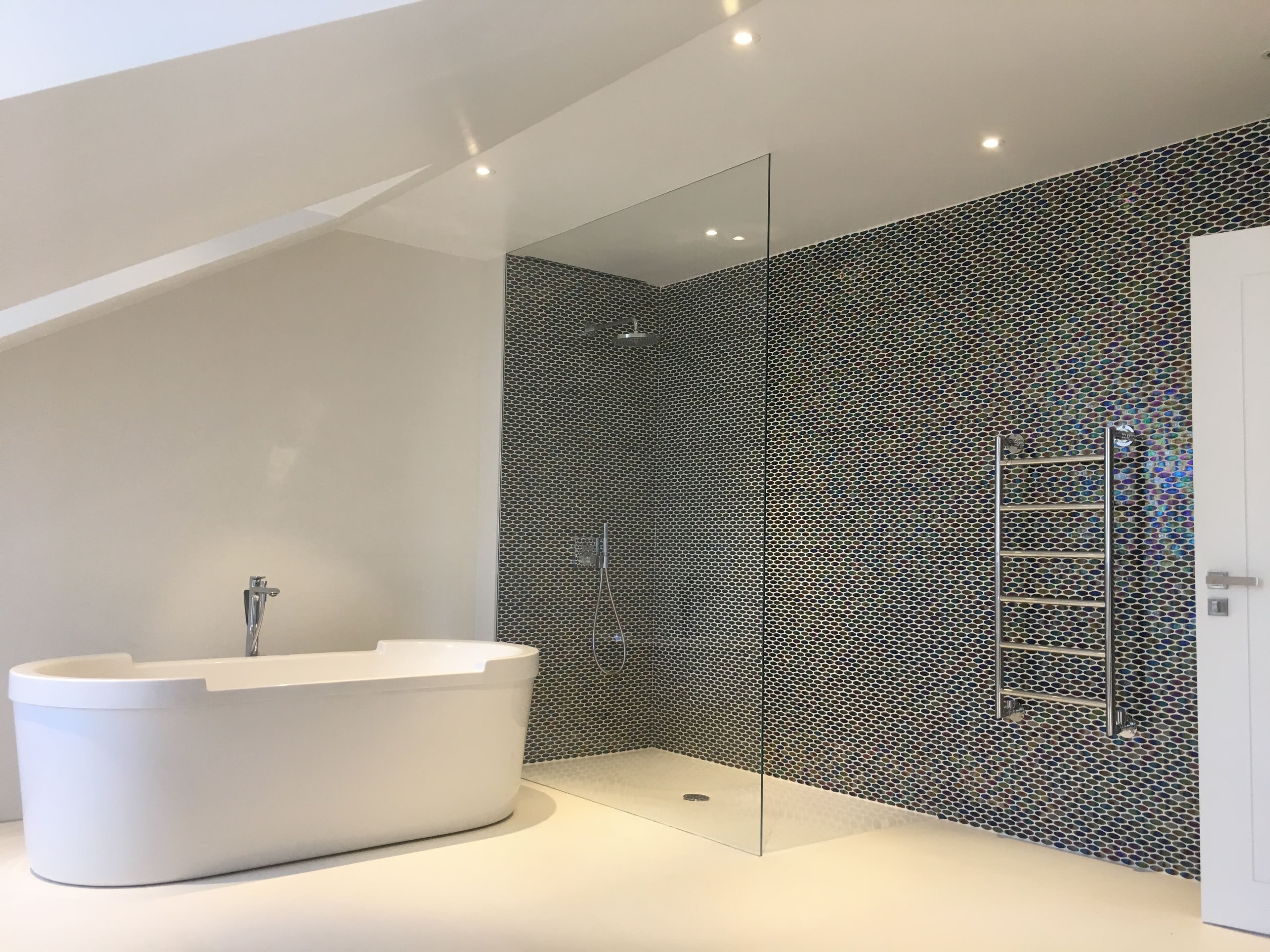 Frameless walk-in shower panel with no visible hardware or frame, mounted beside an iridescent mosaic-tiled wet wall in a contemporary attic bathroom with a freestanding white bath.