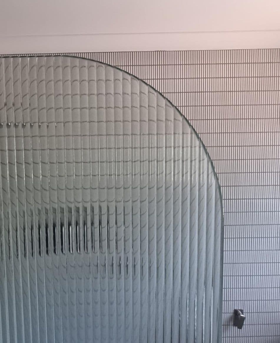 Reeded (fluted) glass panel with a curved arched top edge, shown close-up against a tiled bathroom wall with a small chrome hardware clip visible at the bottom right.