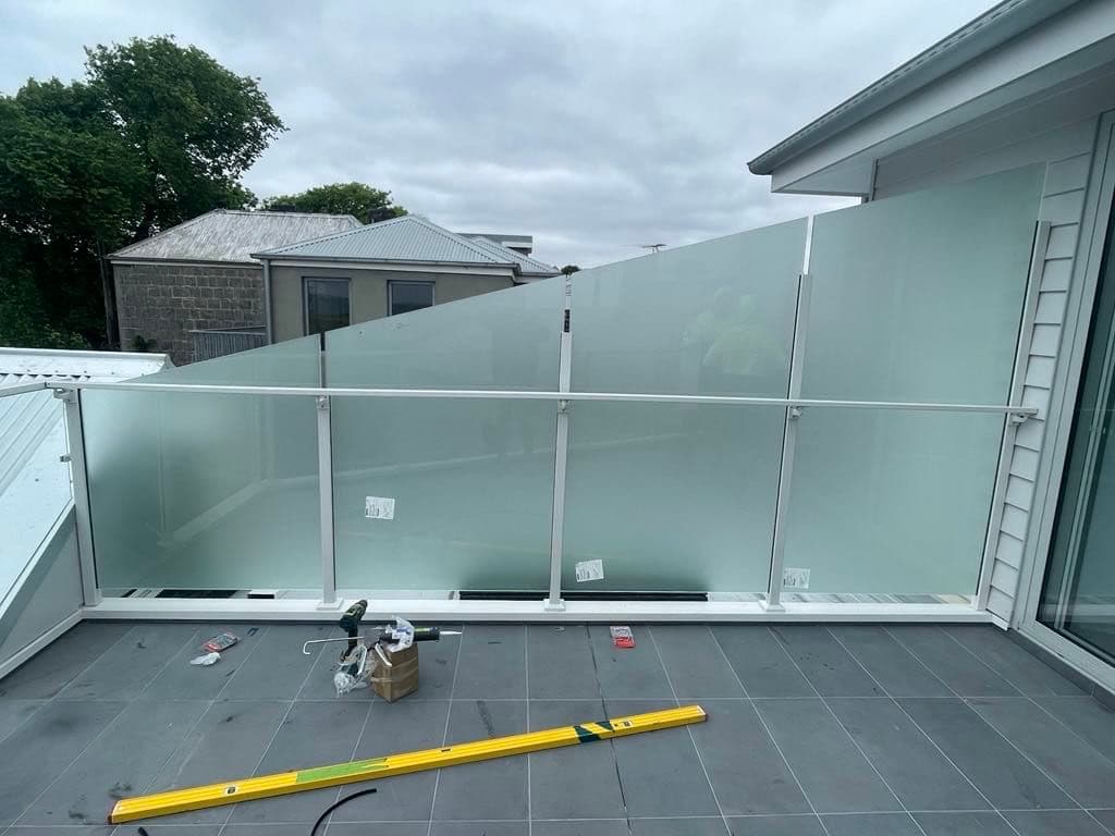 Semi-frameless balustrade with acid etched glass panels, white aluminium top rail and base channel, installed on an elevated tiled balcony during construction.