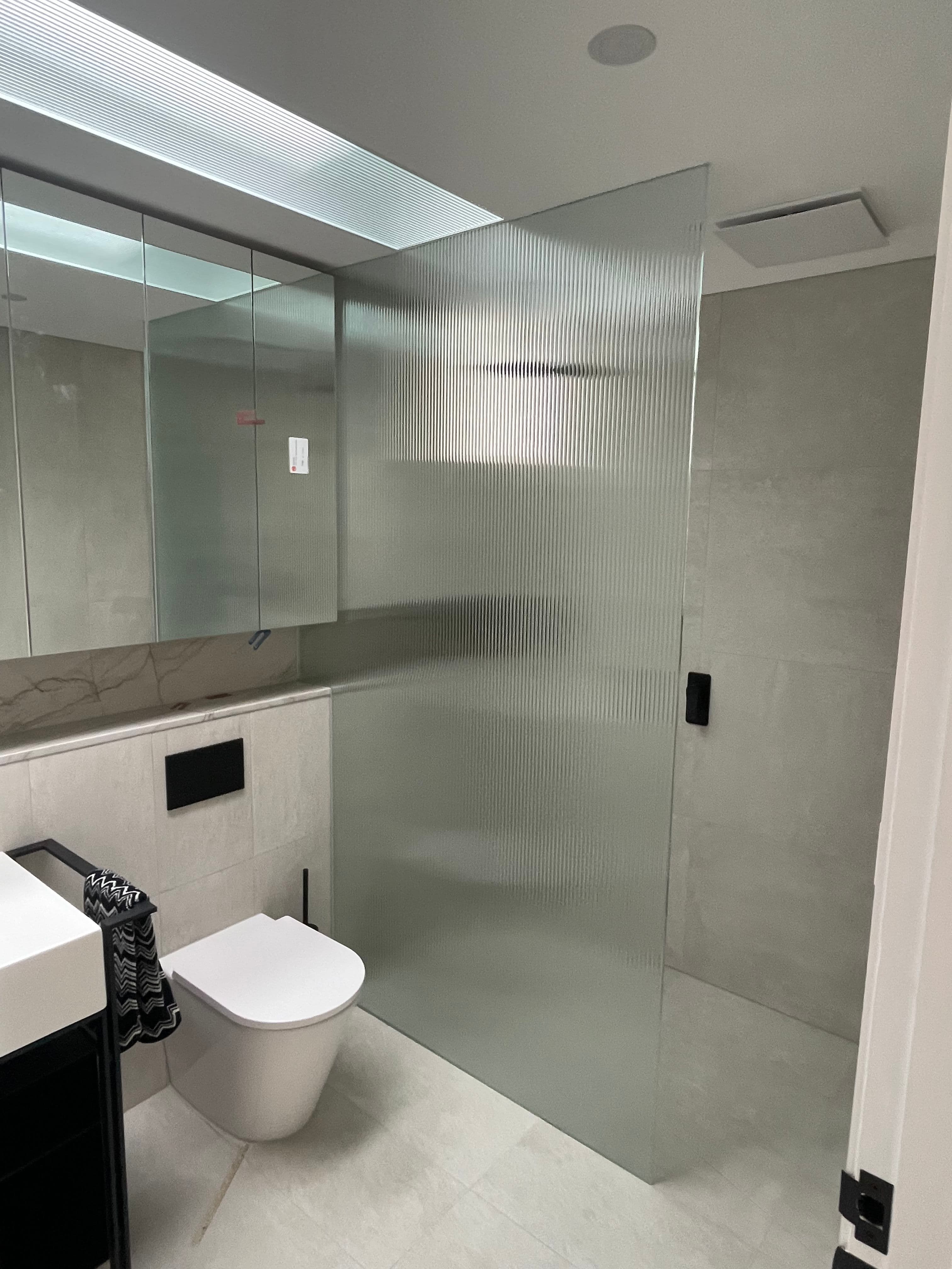 Frameless shower screen featuring reeded fluted glass with a matte black door handle, installed as a fixed panel in a modern tiled ensuite with grey stone-look tiles.