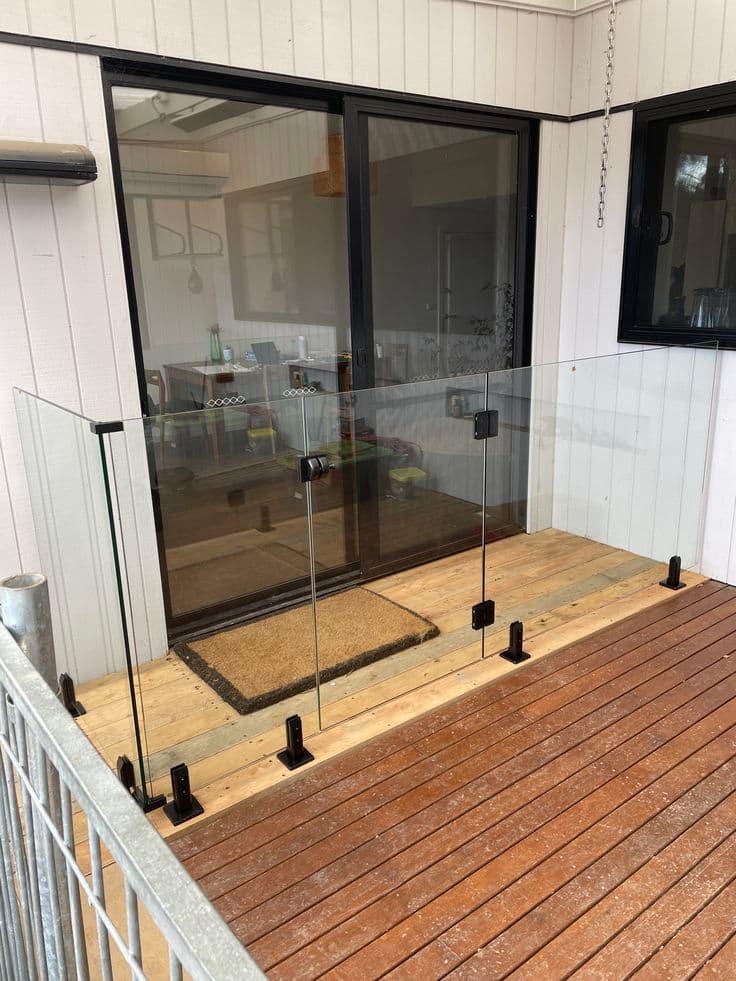 Frameless glass balustrade with matte black square spigots surface-mounted on a timber deck outside a sliding door entry, with no pool visible.