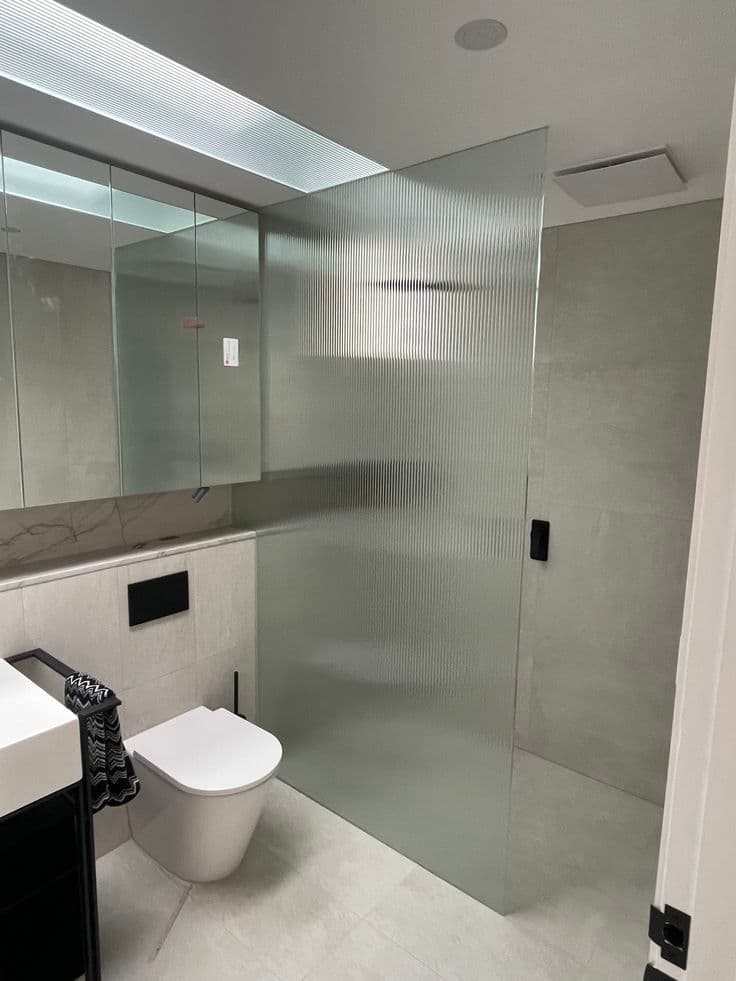 Frameless shower screen in reeded glass with a matte black door handle, installed as a fixed panel in a modern grey-tiled bathroom.