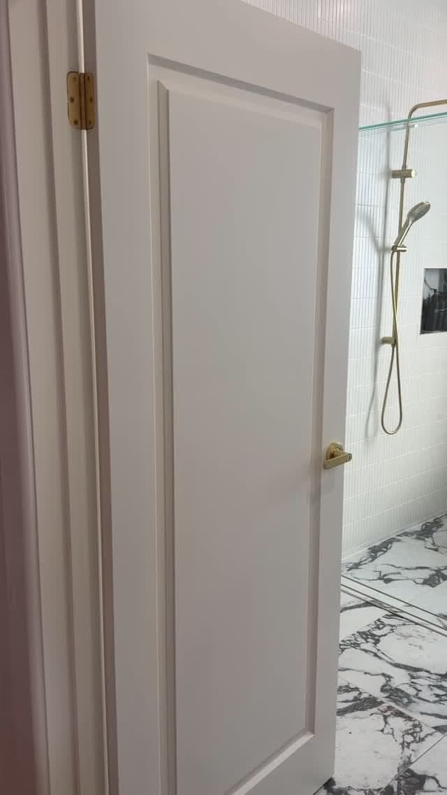 Frameless shower enclosure partially visible behind a white panelled door, with brushed gold shower fittings and clear glass panel in a marble-tiled bathroom.