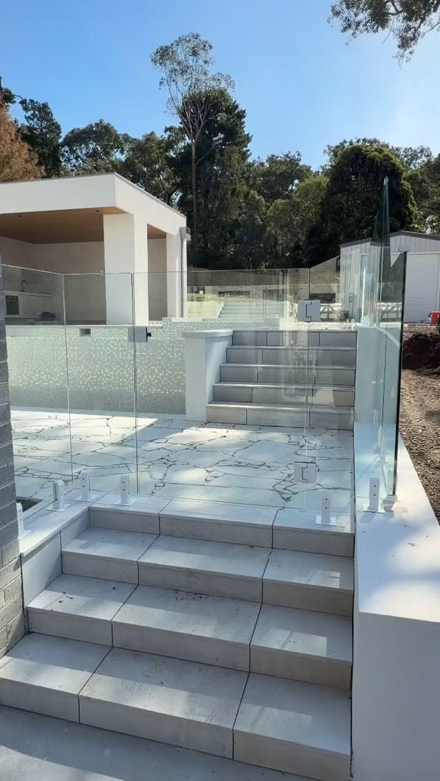 Frameless glass balustrade with brushed chrome spigots installed on a marble-tiled elevated terrace and staircase beside a modern white rendered home exterior.