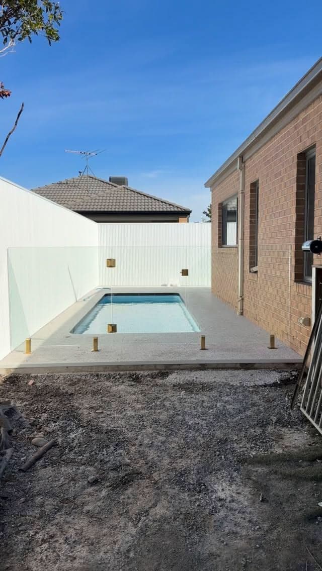 Frameless pool fence with brushed gold square spigots core-drilled into concrete coping surrounding a compact backyard swimming pool beside a brick home.