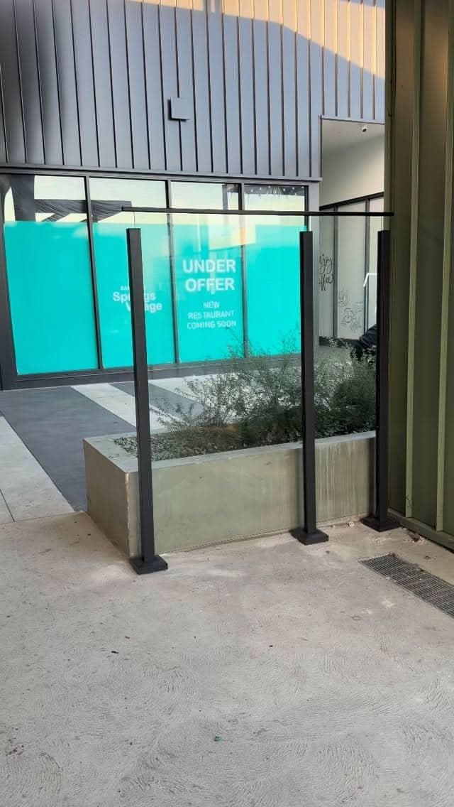 Clear glass balustrade panels with matte black square post hardware mounted on a concrete planter box at a commercial building entrance, no pool visible.