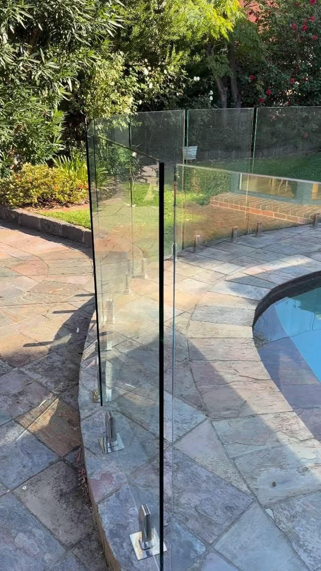 Frameless glass pool fence with chrome spigots core-drilled into stone coping surrounding a backyard swimming pool, with lush garden surroundings.
