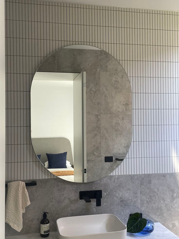 Large round frameless mirror mounted on a tiled bathroom wall above a vessel sink, with matte black tapware and a reflection of an adjoining bedroom.
