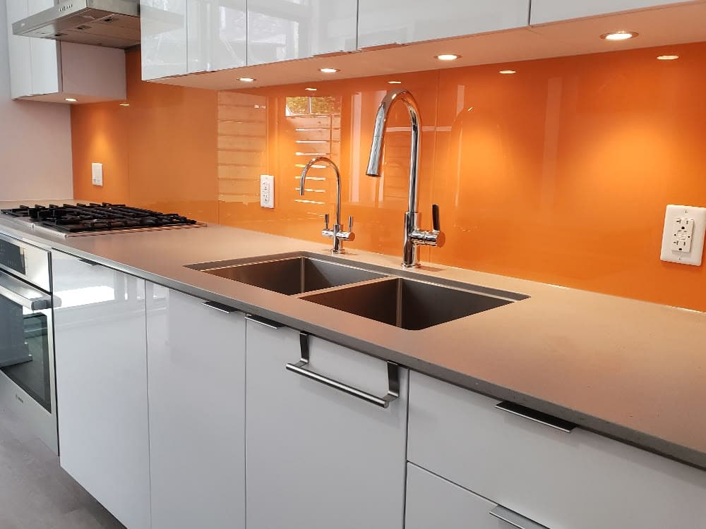 Vibrant orange coloured glass splashback installed behind a gas cooktop and double undermount sink in a modern white kitchen with grey stone benchtop.