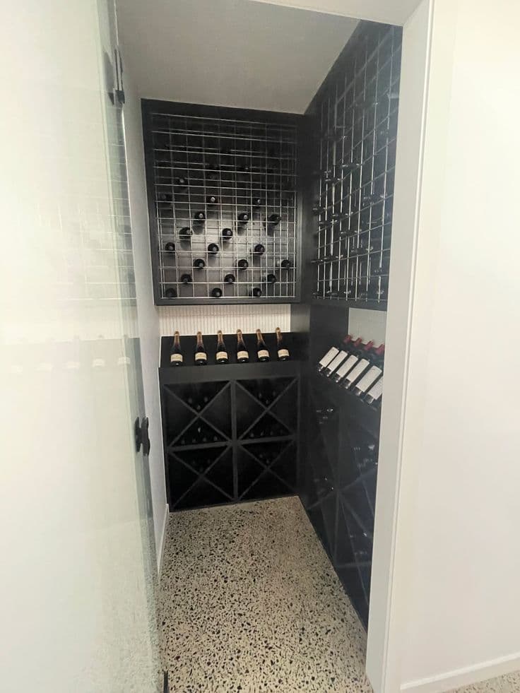 Frameless glass door with matte black hinge hardware providing entry to an interior wine cellar room with terrazzo flooring and black wine racking.