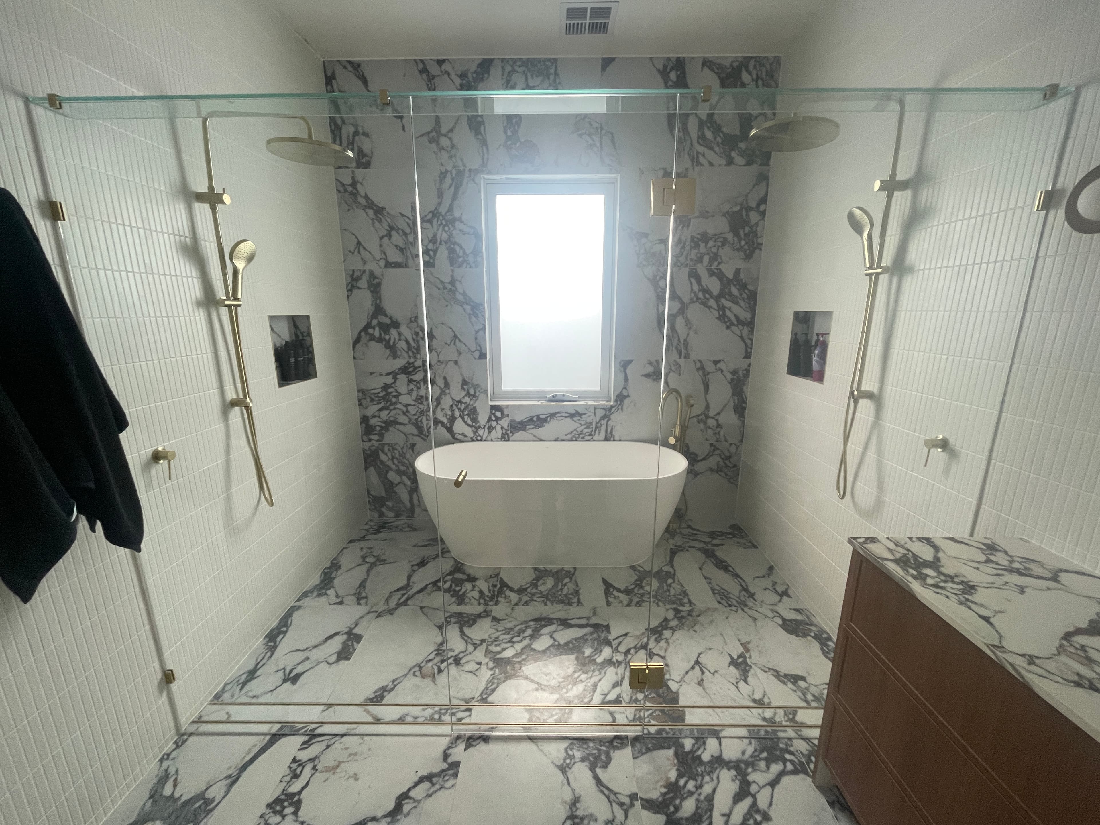 Semi-frameless shower enclosure with brushed gold top rail and side channels in a luxurious marble-tiled bathroom featuring a freestanding bath and dual shower heads.