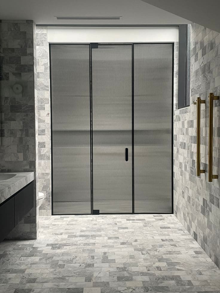 Semi-frameless sliding shower screen with reeded glass panels and matte black aluminium frame channels, installed in a marble-tiled bathroom with brushed gold towel rails.