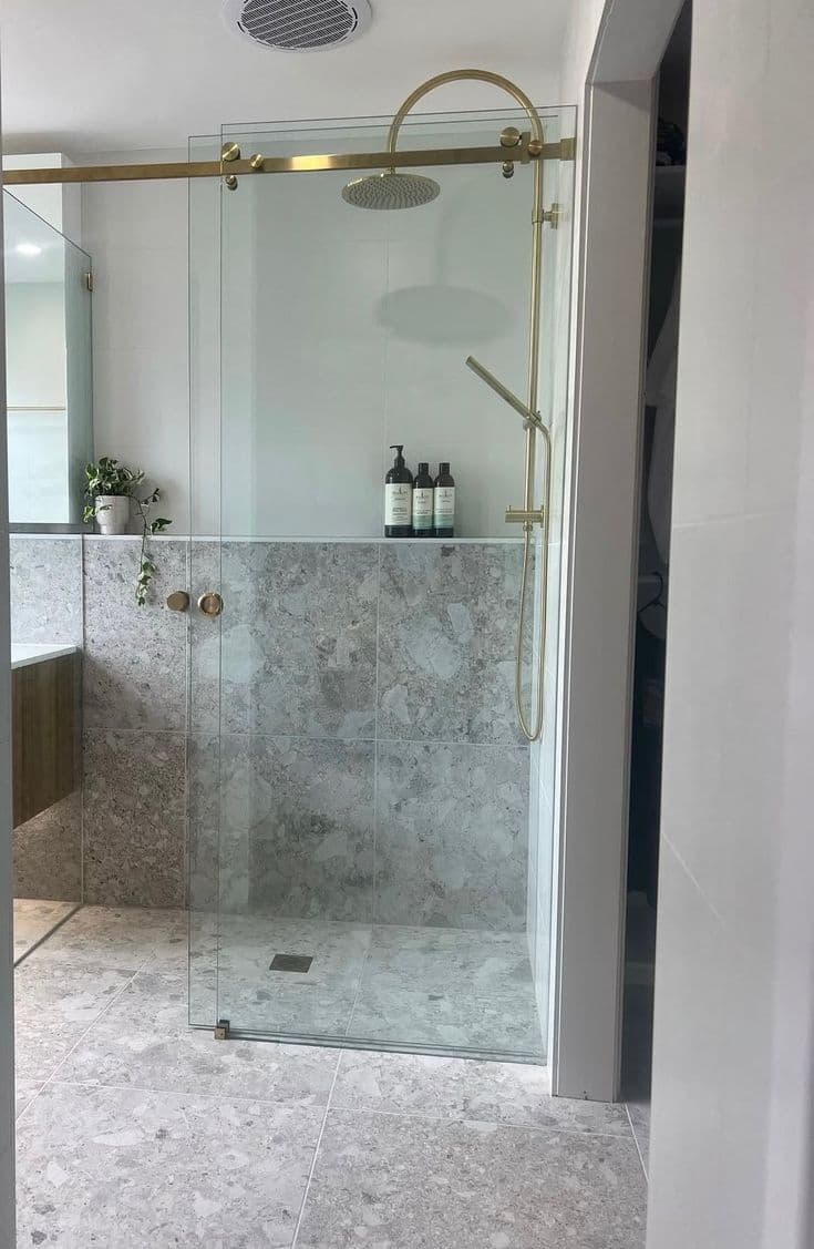 Semi-frameless sliding shower screen with a visible brushed gold top rail and sliding track hardware in a marble-tiled ensuite with matching gold tapware.