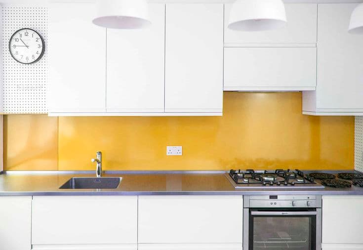 Yellow glass splashback spanning the full width behind a gas cooktop and sink in a modern white kitchen with stainless steel benchtops.