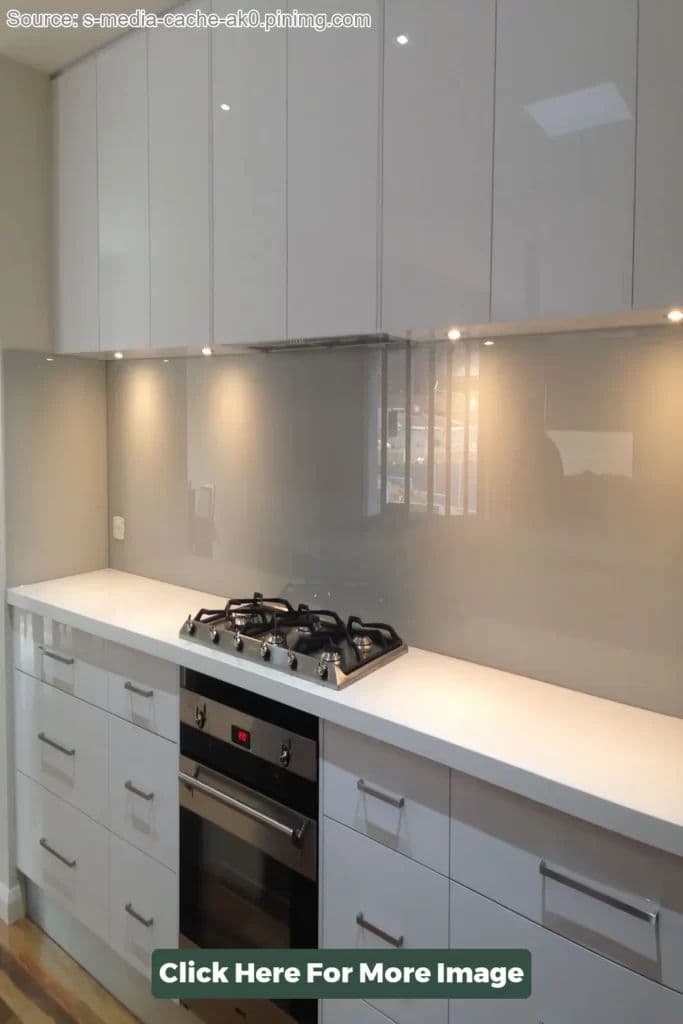 Clear or light grey glass splashback mounted flush on the wall behind a gas cooktop and oven in a modern white gloss kitchen with white stone benchtops.
